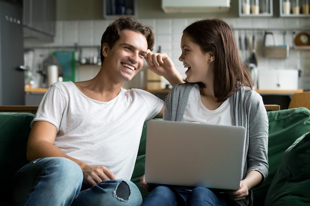 Szczęśliwa millennial para śmia się używać laptop wpólnie na kuchennej kanapie