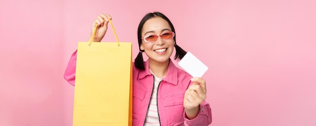 Szczęśliwa młoda azjatycka kobieta pokazująca kartę kredytową na zakupy trzymając torbę kupując na sprzedaż idąc do sho