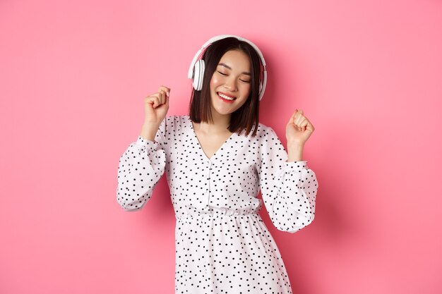 Szczęśliwa młoda azjatycka kobieta tańczy i bawi się, słuchając muzyki w słuchawkach, stojąc na różowym tle. Skopiuj miejsce