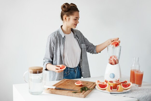 Szczęśliwa młoda dziewczyna miesza zdrowego detox świeżego grapefruitowego smoothie nad biel ścianą.