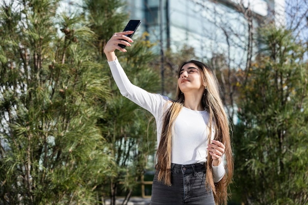 Szczęśliwa młoda dziewczyna robi selfie obok drzewZdjęcie młodej damy na zewnątrz