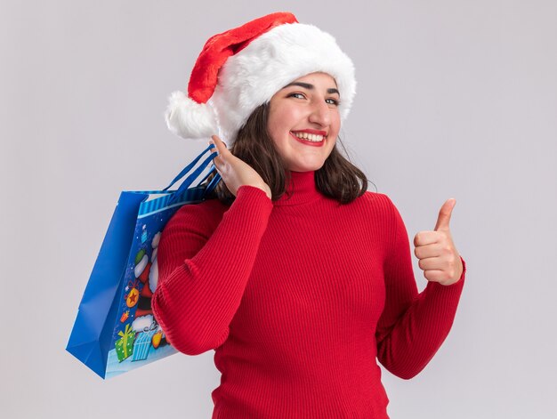 Szczęśliwa młoda dziewczyna w czerwonym swetrze i czapce mikołaja trzymająca kolorową papierową torbę z prezentami bożonarodzeniowymi patrząc na kamerę uśmiechnięta pokazująca kciuki do góry stojąca na białym tle
