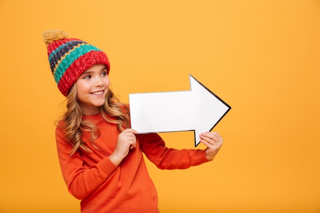 Bezpłatne zdjęcie szczęśliwa młoda dziewczyna w swetrze i kapeluszu patrzeje i wskazuje z papierową strzała daleko od nad pomarańcze