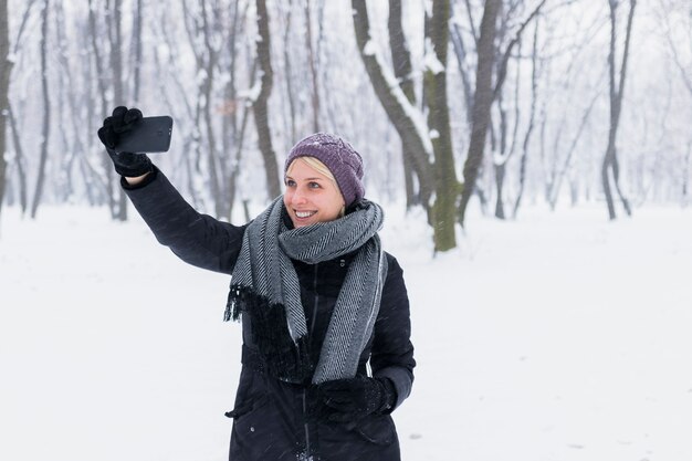 Szczęśliwa młoda kobieta bierze selfie w lesie podczas zimy