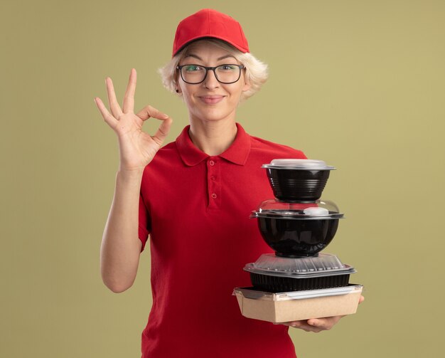 Szczęśliwa młoda kobieta dostawy w czerwonym mundurze i czapce w okularach, trzymając stos opakowań żywności, uśmiechając się pewnie pokazując znak ok stojącego nad zieloną ścianą
