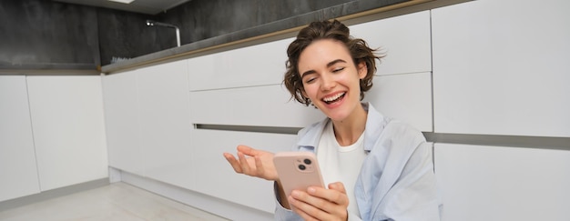 Bezpłatne zdjęcie szczęśliwa młoda kobieta rozmawia przez telefon komórkowy wideo czaty na aplikacji smartfona siedzi na podłodze w domu
