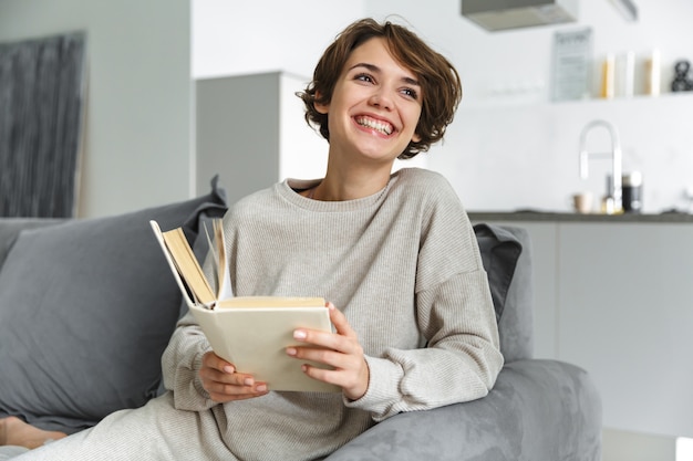 Szczęśliwa młoda kobieta siedzi na kanapie w domu, czytając książkę