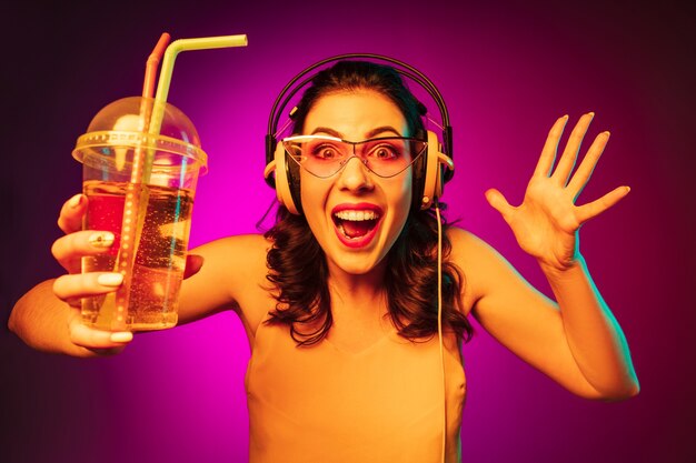 Szczęśliwa młoda kobieta w czerwonych okularach przeciwsłonecznych, picia i słuchania muzyki na modnym różowym neonie
