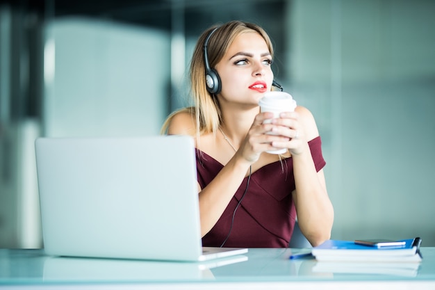 Szczęśliwa młoda kobieta w słuchawkach w call center i picia kawy w biurze.
