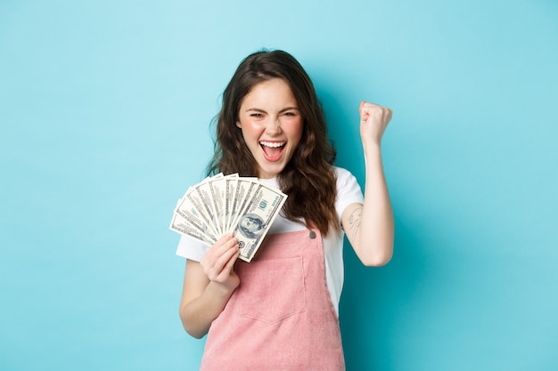Szczęśliwa młoda kobieta wygląda na podekscytowaną, krzycząc z satysfakcji i triumfu, wygrywając pieniądze, trzymając banknoty dolarowe i robiąc pompkę pięścią, stojąc na niebieskim tle