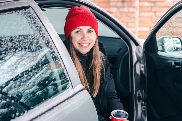 Szczęśliwa młoda kobieta z czerwonym kubkiem termicznym siedzi zimą w samochodzie
