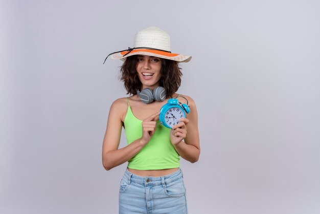 Szczęśliwa młoda kobieta z krótkimi włosami w zielonej bluzce na sobie kapelusz przeciwsłoneczny trzyma niebieski budzik na białym tle