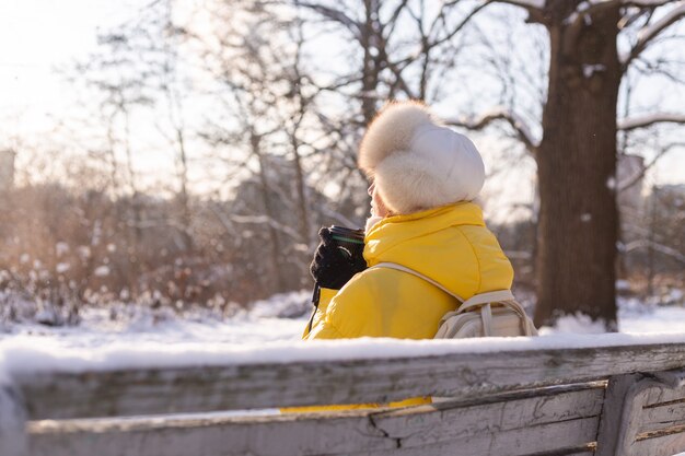 Szczęśliwa młoda kobieta zimą w ciepłych ubraniach w zaśnieżonym parku w słoneczny dzień siedzi na ławkach i sama cieszy się świeżym powietrzem i kawą