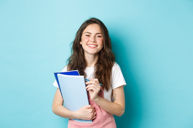 Szczęśliwa młoda studentka trzyma zeszyty z kursów i uśmiecha się do kamery, stojąc w wiosennych ubraniach na niebieskim tle.