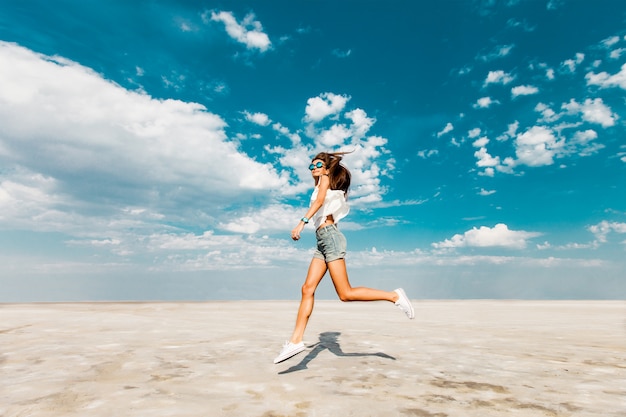 Szczęśliwa młoda świeża szczupła dziewczyna lekkoatletycznego biegnie wzdłuż plaży w modnych dżinsowych szortach i białych trampkach. Błękitne niebo w chmurach, letni słoneczny nastrój.