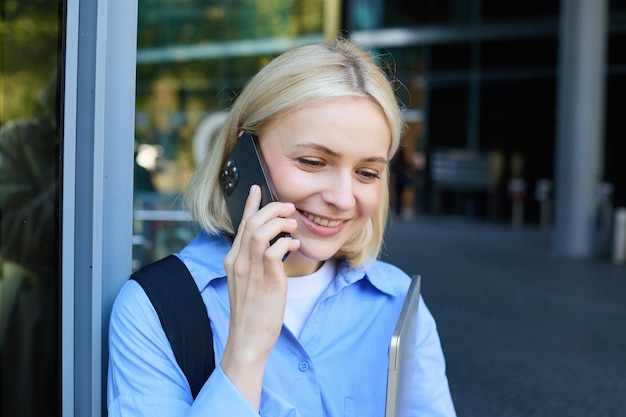 Bezpłatne zdjęcie szczęśliwa modelka blondynka rozmawiająca przez telefon komórkowy rozmawiająca z kimś stojącym w pobliżu biura