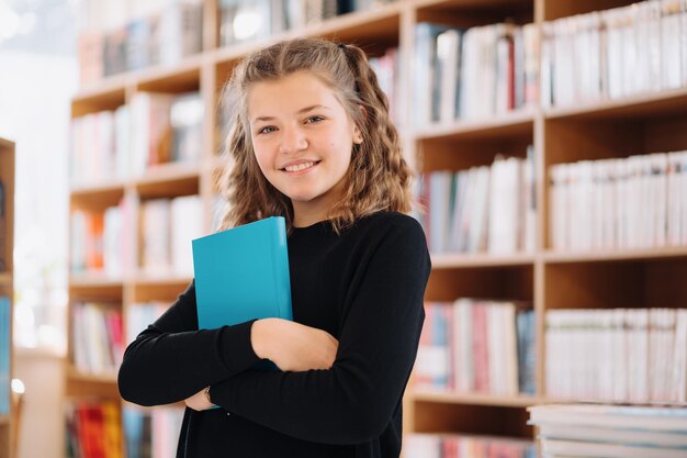 Szczęśliwa nastoletnia dziewczyna lub uśmiechnięty uczeń trzymający niebieską książkę z miejscem na kopię wśród wielu książek w bibliotece - koncepcja ludzie, wiedza, edukacja i szkoła