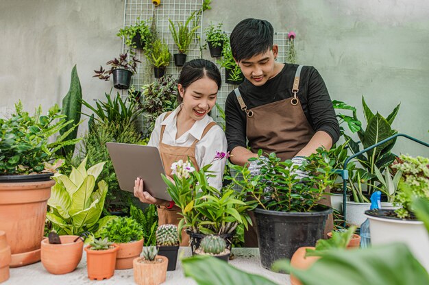 Szczęśliwa para młodych ogrodników z Azji w fartuchach korzysta ze sprzętu ogrodowego i laptopa, aby się opiekować