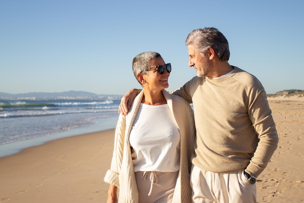 Szczęśliwa para starszych spacerując brzegiem morza, spędzając razem czas na wakacjach. Siwy mężczyzna, obejmując uśmiechniętą krótkowłosą kobietę w okularach przeciwsłonecznych. Pojęcie związku, emerytury, stylu życia