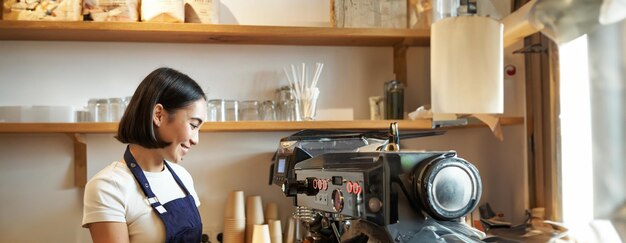 Szczęśliwa, piękna Azjatka, barista w fartuchu, pracuje z ekspresem do kawy za ladą.