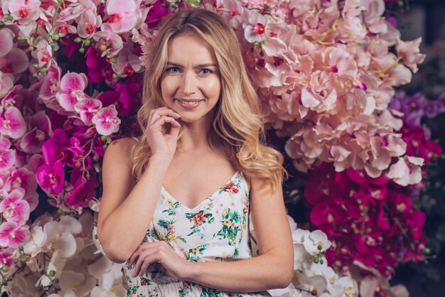 Szczęśliwa piękna blondynki młoda kobieta z ręką na podbródek pozyci przed storczykowymi kwiatami