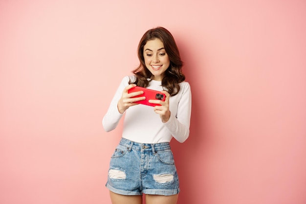 Szczęśliwa piękna dziewczyna grająca w mobilną grę wideo, trzymająca smartfon poziomo, oglądająca na telefonie komórkowym z podekscytowaną twarzą, różowym tle