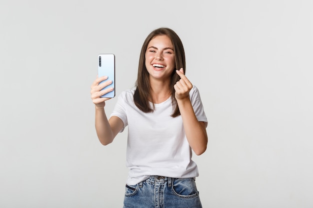 Szczęśliwa piękna młoda kobieta pokazuje gest serca i bierze selfie na smartfonie, śmiejąc się beztrosko.