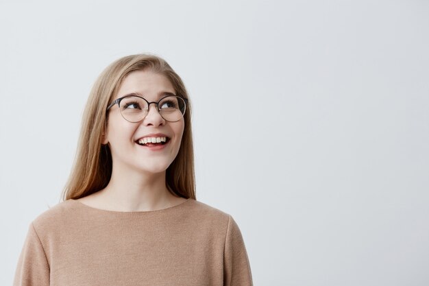 Szczęśliwa podekscytowana zdziwiona modelka z prostymi blond włosami w okularach, będąc z czegoś zadowolona, nie może uwierzyć w swój sukces, cieszę się z pochwały, pozuje na szarym tle studia