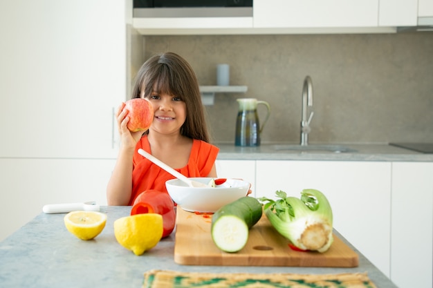 Szczęśliwa pozytywna dziewczyna stojąca przy blacie kuchennym z ciętych świeżych warzyw, trzymając i pokazując jabłko, uśmiechając się, patrząc na kamery. Pojęcie zdrowego odżywiania