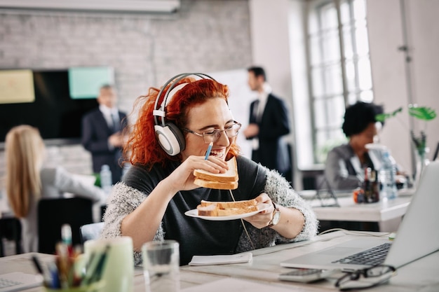 Szczęśliwa przypadkowa bizneswoman bawi się i śmieje podczas jedzenia kanapki podczas przerwy w pracy W tle są ludzie