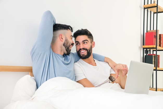 Szczęśliwa przystojna homoseksualna para siedzi wpólnie na łóżku z laptopem