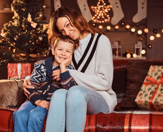 Szczęśliwa rodzina. Mama przytula swojego słodkiego małego chłopca siedząc na kanapie w urządzonym pokoju w czasie świąt Bożego Narodzenia.