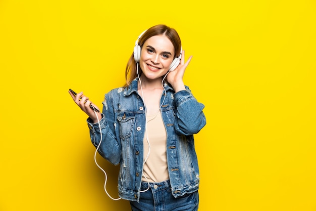 Szczęśliwa rozochocona kobieta jest ubranym hełmofony słucha muzyka od smartphone studia strzelającego odizolowywającym na kolor żółty ścianie