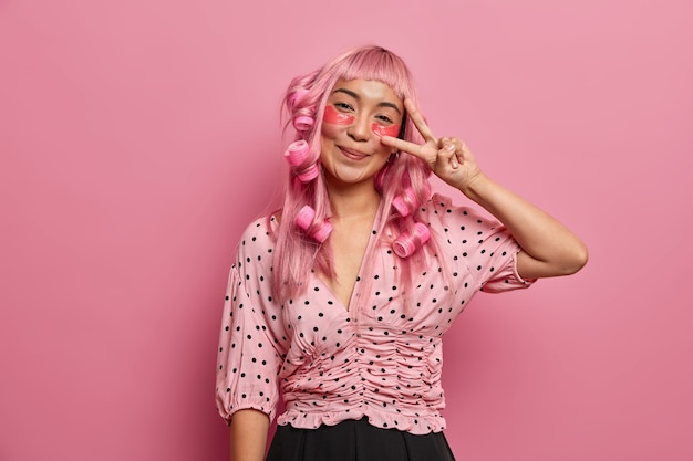 Bezpłatne zdjęcie szczęśliwa różowowłosa dziewczyna nakłada plastry na oczy, aby zmniejszyć cienie pod oczami, robi znak pokoju, nosi lokówki, aby uzyskać idealne loki
