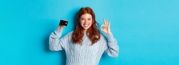 Bezpłatne zdjęcie szczęśliwa ruda dziewczyna w swetrze pokazująca kartę kredytową i dobry znak polecający ofertę bankową na stojąco