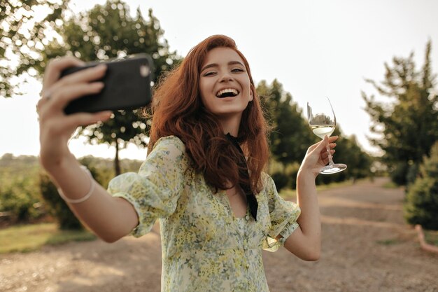 Szczęśliwa rudowłosa kobieta z czarnym bandażem na szyi w zielonej sukience, śmiejąc się, robiąc selfie i pozując ze szklanką z szampanem na świeżym powietrzu