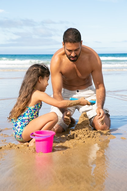 Szczęśliwa śliczna mała dziewczynka i jej tata budują zamek z piasku na plaży, siedząc na mokrym piasku, ciesząc się wakacjami