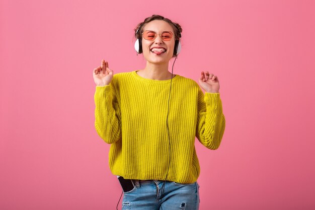 Szczęśliwa śmieszna kobieta słuchająca muzyki w słuchawkach ubrana w strój kolorowy hipster na białym tle na różowej ścianie, ubrany w żółty sweter i okulary przeciwsłoneczne, bawiąc się pokazując język