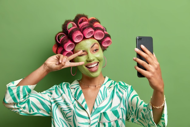 Szczęśliwa śmieszna Kobieta Sprawia, że Selfie Kształtuje Znak Zwycięstwa W Smartfonie Aparat Uśmiecha Się Szeroko Cieszy Zabiegi Na Twarz Stosuje Wałki Do Włosów Ubrane W Zwykłe Domowe Ubrania Na Białym Tle Nad Zieloną ścianą