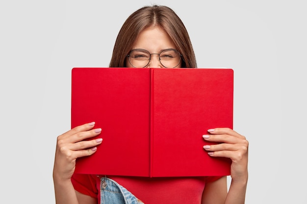 Szczęśliwa studentka śmieje się pozytywnie, nosi okrągłe okulary, chowa się za czerwoną książeczką, uśmiecha się, czytając coś śmiesznego, pozuje przy białej ścianie. Koncepcja ludzi, młodzieży, edukacji i czytania