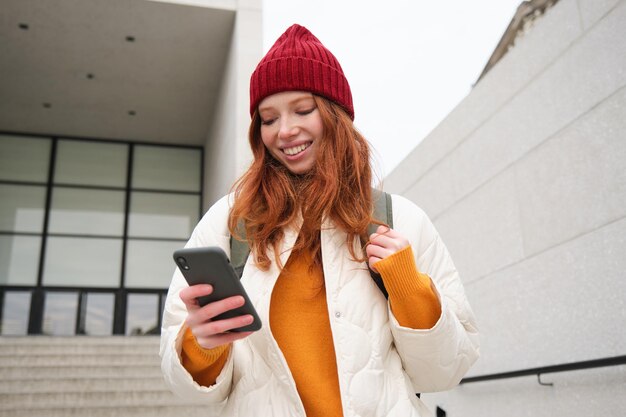 Szczęśliwa studentka w czerwonym kapeluszu trzyma smartfona, turysta patrzy na aplikację mapy na swoim telefonie, odkrywa zabytki