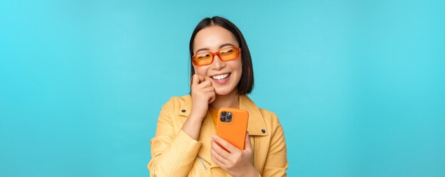 Szczęśliwa stylowa azjatycka dziewczyna używająca smartfona i śmiejąca się, uśmiechając się do kamery stojącej na niebieskim tle