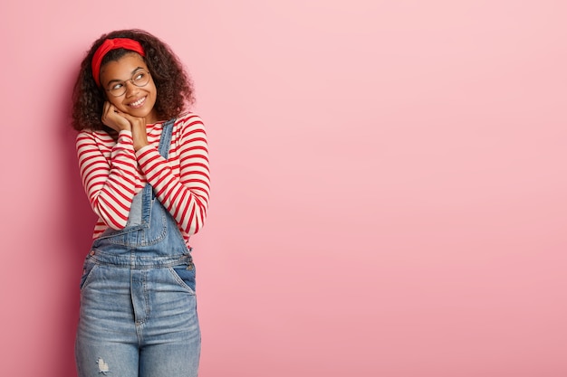 Szczęśliwa Tysiącletnia Afroamerykańska Dziewczyna Wygląda Radośnie Na Bok, Nosi Czerwoną Opaskę, Sweter W Paski I Dżinsowy Kombinezon