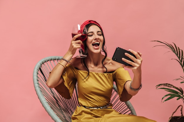 Szczęśliwa Urocza Kobieta Z Falującymi Ciemnymi Włosami W Czerwonej Chustce I Kolczykach Pozuje Ze Smartfonem I Trzyma Kieliszek Wina Na Różowej ścianie