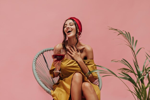 Szczęśliwa urocza kobieta z kolczykami z czerwoną opaską i modnym jasnym strojem, śmiejąca się z zamkniętymi oczami i pozująca z winem