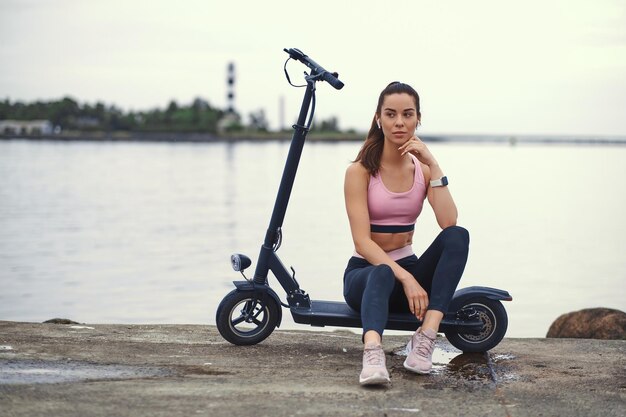 Szczęśliwa wesoła kobieta w odzieży sportowej relaksuje się na swoim skuterze elektrycznym nad brzegiem morza.