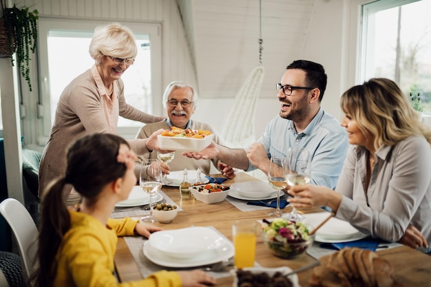 Szczęśliwa wielopokoleniowa rodzina je razem obiad w domu Starsza kobieta przynosi jedzenie na stół