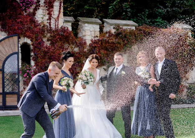 Szczęśliwe druhny, najlepsi mężczyźni i para weselna świętują dzień ślubu na zewnątrz, wylewając szampana