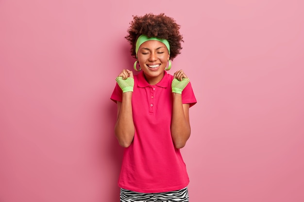 Szczęśliwe emocje i uczucia. Uśmiechnięta afroamerykańska dziewczyna w różowej koszulce, sportowych rękawiczkach i opasce, zaciska z radości pięści, czuje smak zwycięstwa, świętuje zwycięstwo w konkursie