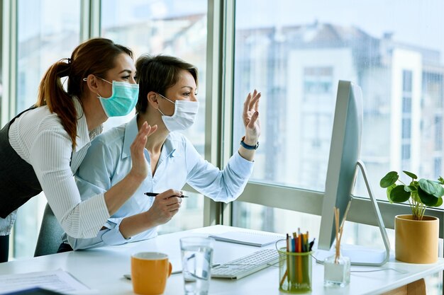 Szczęśliwe kobiety biznesu z machającymi maskami podczas prowadzenia wideorozmowy na komputerze stacjonarnym w biurze podczas epidemii wirusa